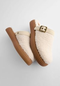 Sabots crème avec un dessus en shearling pelucheux, une sangle beige avec boucle en laiton, et une semelle en caoutchouc marron. Texture douce et design casual.