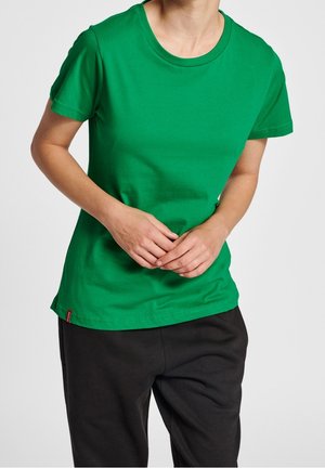 Person trägt ein schlichtes grünes T-Shirt mit kurzen Ärmeln und weite schwarze Hosen, steht mit verschränkten Händen vor dem Körper.