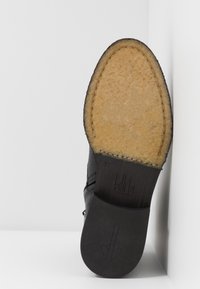 Suela de zapato de cuero negro con superficie texturizada, plataforma intermedia de color marrón y borde cosido. Presenta el nombre de la marca y marcas de tamaño en relieve.