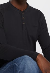 Svart långärmad henley-tröja med treknappsplacket och en liten logo. Släta tyger med en avslappnad passform. Parat med denimjeans.