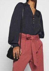 Marinebluse med puffede ærmer, v-hals og tre knapper; stylet med højtsiddende lyserøde bukser med en bundet bælt; sort crossbody taske.