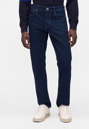 Homme portant un jean bleu foncé et des baskets blanches, debout devant un fond clair uni, mains détendues le long du corps.