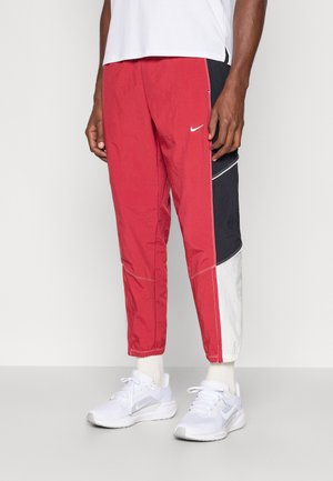 Mężczyzna ubrany w czerwone, czarne i białe spodnie dresowe Nike, białą koszulkę i białe sneakersy Nike, stoi na jednolitym tle.