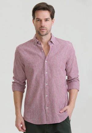 Mand iført en rød og hvid lodret stribet skjorte med opkørte ærmer og mørke bukser, står foran en enkel lys baggrund.