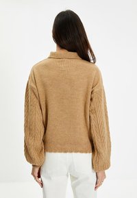 Suéter de punto marrón con un corte corto, que presenta un dobladillo acanalado y un patrón de cable texturizado en las mangas voluminosas. Pantalones blancos completan el look.