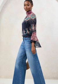 Bluza z dolgimi rokavi v mornarsko modri barvi z vzorcem paisley, visokim ovratnikom in zvončastimi rokavi, v kombinaciji s širokimi modrimi džinsami; gladka tekstura in zračen dizajn.