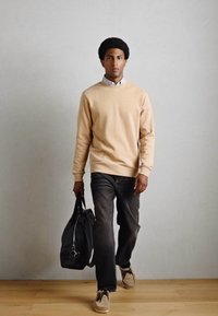 Pull en coton beige, chemise col bleu clair en dessous, jean foncé, portant un sac en cuir noir, debout sur un sol en bois contre un mur gris.