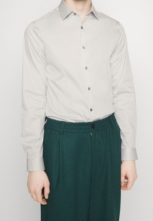 Camicia leggera grigia a maniche lunghe con colletto a punta, bottoni frontali e texture liscia, abbinata a pantaloni verdi a fantasia e un bottone nero.