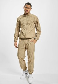 Lys beige treningsdrakt med hettejakke som har glidelås og elastiske mansjetter, samt matchende bukser med elastisk liv. Kombinert med hvite sneakers.