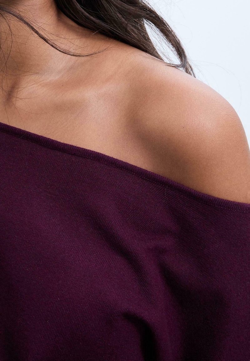 Épaule et haut de la poitrine d'une femme visibles, portant un haut violet foncé décolleté aux épaules avec des cheveux foncés lâches qui tombent.