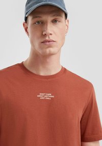 Bruine katoenen t-shirt met korte mouwen en een ronde hals. De tekst op de voorkant luidt: "GEEN ZORG OVER IETS, GEWOON RELAXEN."