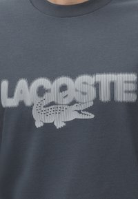 Σκούρο γκρι πουκάμισο με τη λέξη "LACOSTE" σε μεγάλες λευκές κάθετες γραμμές, με ένα λευκό λογότυπο κροκοδείλου ενσωματωμένο στο κείμενο.