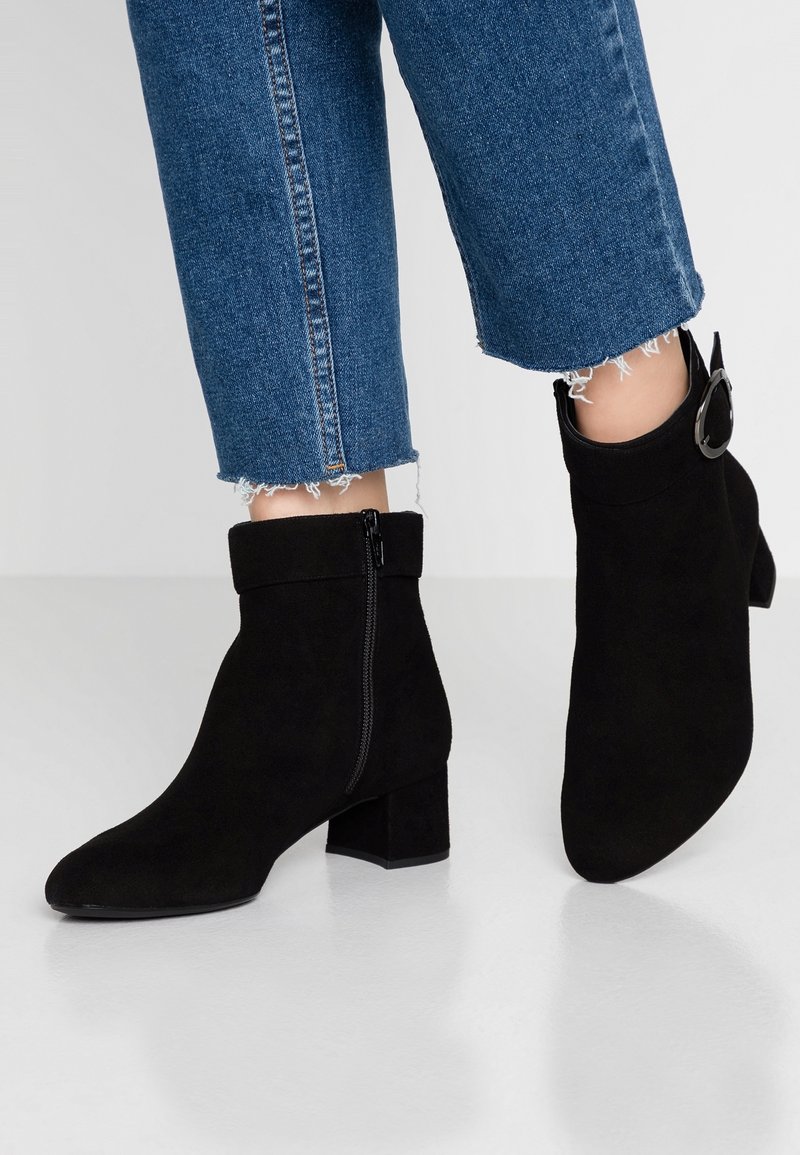 Bottines en daim noir à talon bloc, avec une fermeture éclair latérale et un revers plié. Portées avec un denim bleu raccourci, aux ourlets effilochés.