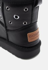 Botte en cuir noir avec des œillets en métal argenté, un col en tissu texturé et une étiquette en brun avec le logo "GIOSEPPO" et l'inscription "HAVE A NICE DAY".