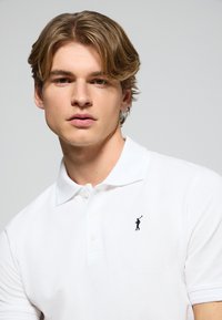 Polo bianco dal design elegante, con una patta a tre bottoni e un piccolo logo ricamato di un golfista in nero sul lato sinistro del petto.