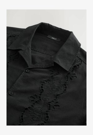 Svart skjorta med broderad blommönster längs knäppningen. Har en spridd krage och knappstängning, tillverkad av ett strukturerat tyg.