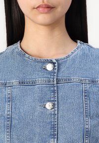 Denim jakna, svetlo modra, z okroglim izrezom, dvema sprednjima gumboma in vidnimi šivi. Gladka tekstura z rahlim bledenjem.