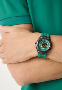Reloj analógico verde con una caja hexagonal negra, esfera multicolor y una correa de silicona verde texturizada. Lleva puesto en una muñeca contra una camisa verde.