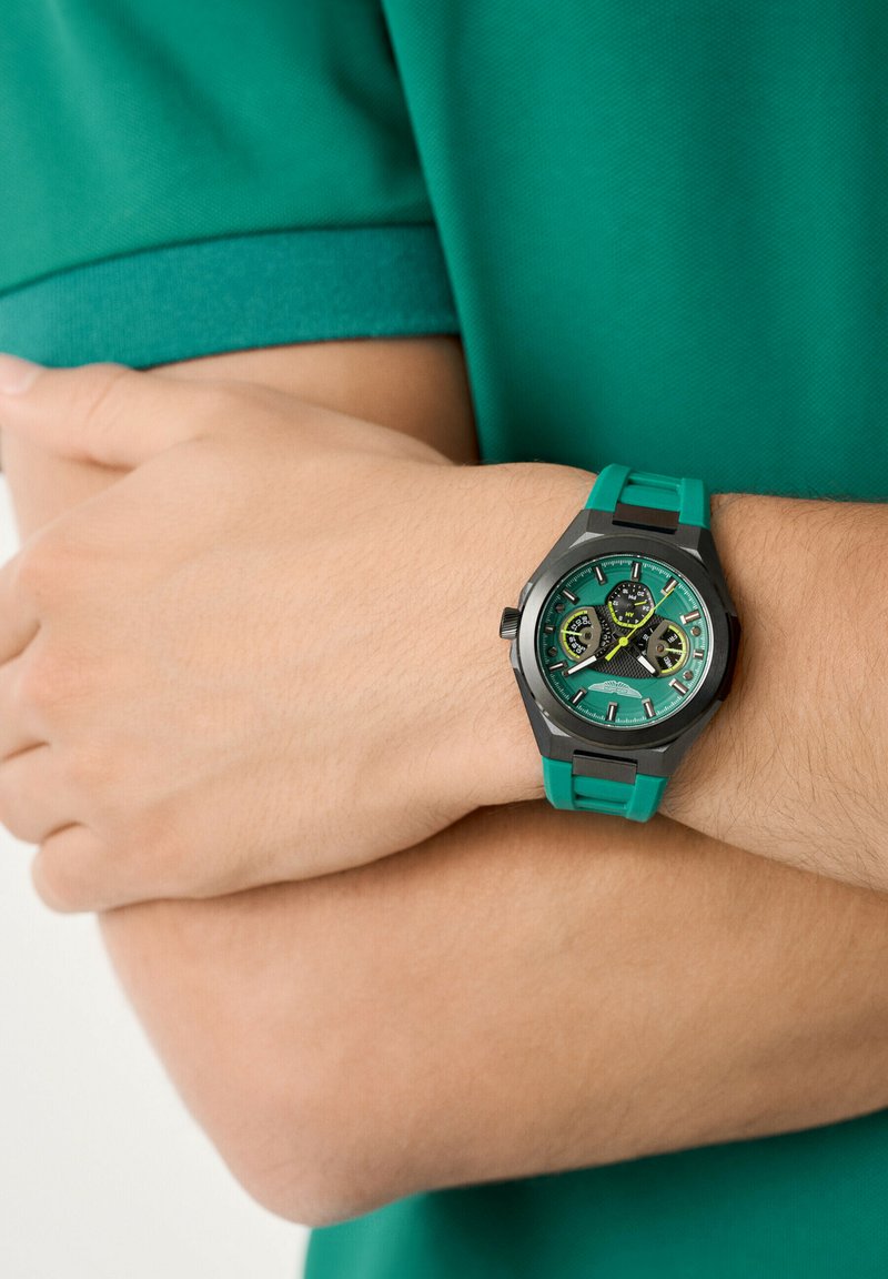 Reloj analógico verde con una caja hexagonal negra, esfera multicolor y una correa de silicona verde texturizada. Lleva puesto en una muñeca contra una camisa verde.
