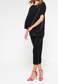 Žena v černém texturovaném topu a kalhotách s černou kabelkou přes rameno, stojící na červených vysokých podpatcích na bílém pozadí.