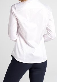 Chemise blanche à manches longues avec une texture lisse, design ajusté et un léger arrondi à l'ourlet, associée à un pantalon ajusté bleu marine.