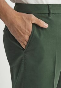 Mörkgröna byxor med en slät textur, som har sidofickor och en åtsittande midja, delvis visade med en hand i fickan.