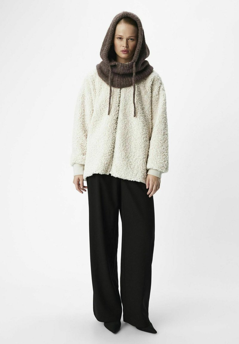 Pull en polaire beige crémeux et moelleux avec capuche, présentant un col tricoté marron et des cordons de serrage, assorti à un pantalon large noir.