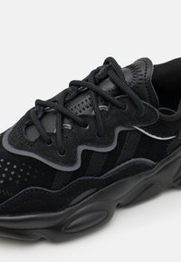 Black athletic shoes featuring a mix of suede and leather, textured panels, wavy design elements, and cushioned sole with circular accents.