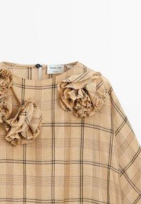 Beige geruite top met ruches bij de schouder, met geometrische vormen in een lichte stof. Het label leest "Massimo Dutti Studio."