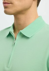 Zelená polo tričko s texturovaným límcem, které má zapínání na zip. Hladká látka s mírným leskem, těsně padnoucí design a jednobarevné provedení.