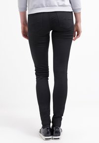 Jean skinny noir avec une texture lisse, design à cinq poches et un discret détail de couture à l'arrière, associés à des baskets noires.