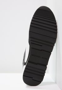 Černá podešev tenisek s vroubkovanou texturou. Obsahuje bílý akcent s logem "MICHAEL KORS." Materiál vypadá jako guma s hladkým povrchem.