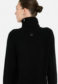 Maglione a collo alto di colore nero in materiale a costine, caratterizzato da un colletto alto e un piccolo dettaglio con logo sulla parte posteriore, con maniche lunghe aderenti.