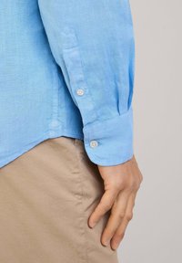 Camicia in lino azzurro chiaro con polsini a bottone e orlo spaccato. Mano appoggiata su pantaloni beige, che mostrano una finitura pulita e testurizzata.