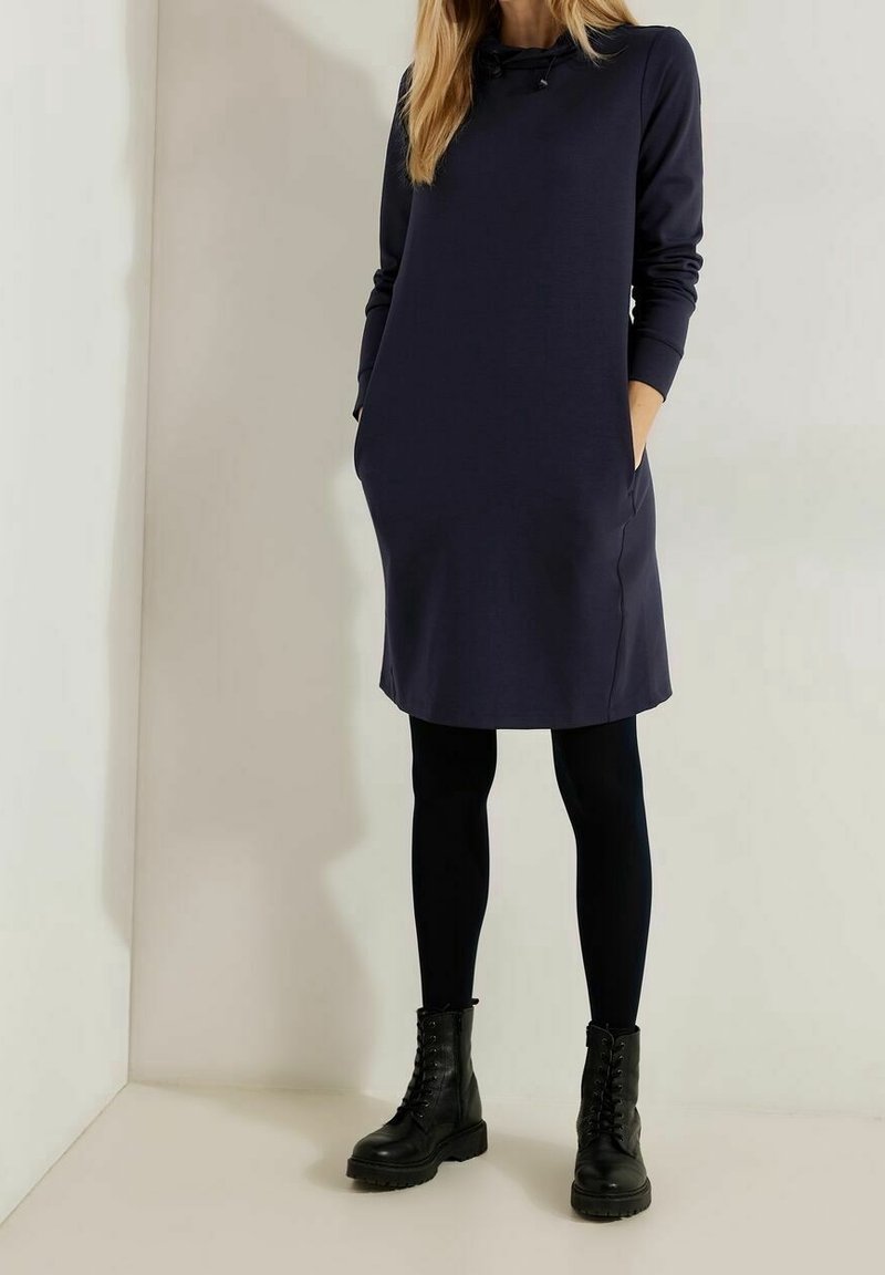 Femme portant une robe à capuche bleu marine à manches longues avec des poches, des leggings noirs et des bottes de combat noires à lacets, debout contre un mur uni.