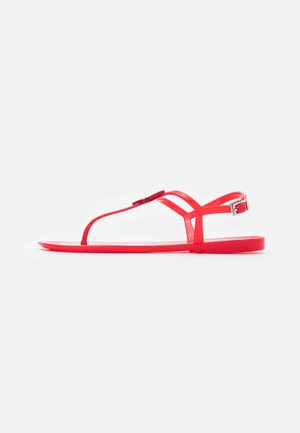 Rød flad sandal med tynde remme, en sølvspænde og en tårem, vist i sideprofil på hvid baggrund.