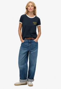 Vrouw staat met handen in de zakken, draagt een marineblauw t-shirt met witte biezen, brede blauwe jeans met opgerolde broekspijpen, een bruine riem en beige sneakers.