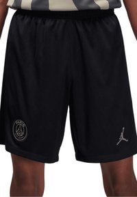 Shorts de sport noirs avec le logo du Paris Saint-Germain sur la jambe gauche et le logo Jumpman sur la jambe droite, portés avec une chemise rayée.