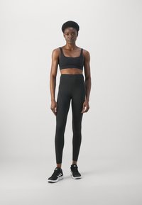 Fekete sportos crop top vékony pántokkal, magas derekú fekete leggingsszel párosítva. A modellen fekete sneakers van, fehér díszítéssel.