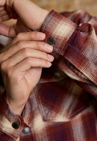 Hands fastening the cuff of a red and beige plaid shirt with a black snap button.