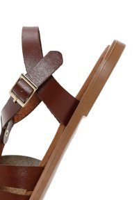 Sandal in pelle marrone con cinturino regolabile e fibbia metallica, dotata di suola piatta color tan e plantare testurizzato. Design minimalista e stile aperto.