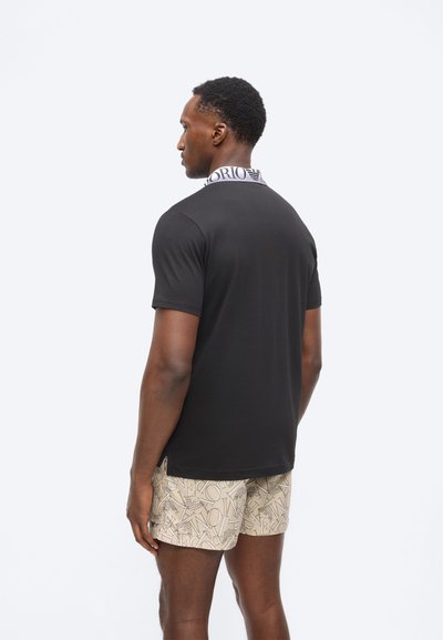 Άνδρας που κοιτάζει προς τα αριστερά, φοράει μαύρο πουκάμισο με κοντά μανίκια και λευκό λογότυπο στο γιακά, καθώς και μπεζ σορτς με αφηρημένα μοτίβα γραμμάτων.