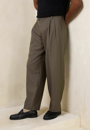 Person wearing loose, pleated brown trousers and black loafers, standing with hands behind back against a light beige wall.