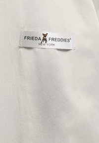 Witte stof met een gladde textuur heeft een label met de tekst "FRIEDA FREDDIES" in zilver, met een klein bruin dierenlogo.