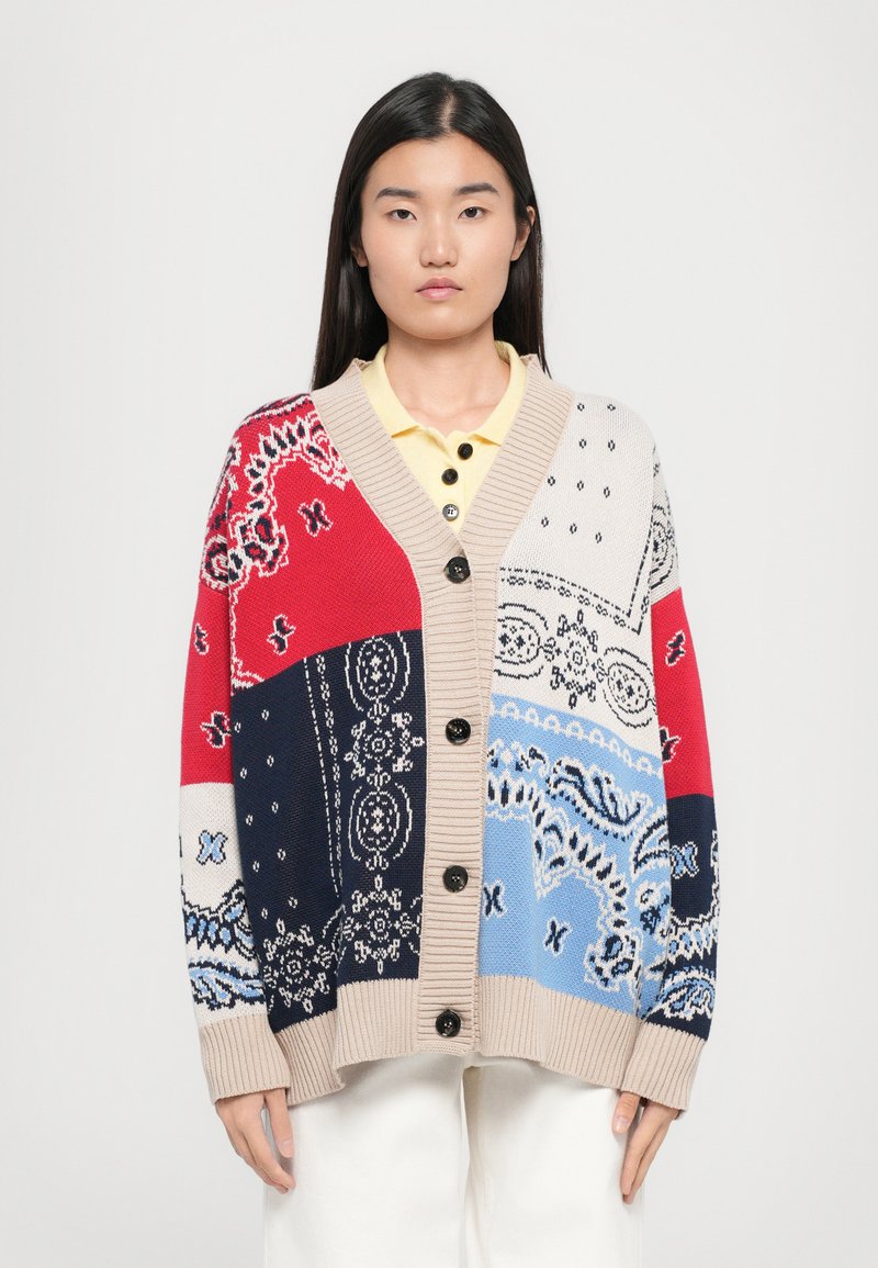 Cardigan en tricot avec un design en blocs de couleur rouge, bleu, beige et des accents à motifs. Présente un col en V, une fermeture à boutons à l'avant et des poignets côtelés.