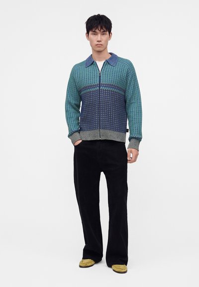 Jeune homme debout portant un pull zippé vert et bleu, un pantalon noir et des chaussures jaunes sur un fond blanc uni.