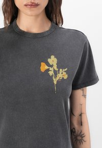 T-shirt in cotone grigio con una piccola stampa floreale gialla e arancione sul lato sinistro del petto, collo rotondo e maniche corte.