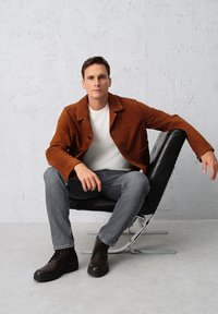 Brązowa zamszowa kurtka, biała koszulka, szare dżinsy oraz ciemnobrązowe sznurowane buty. Siedzi na czarnym krześle na minimalistycznym tle.