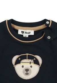 Schwarzes Baumwoll-T-Shirt mit einem strukturierten, gestickten Bärenkopf mit Mütze, beigen Akzenten und Druckknöpfen an der Schulter. Markenetikett sichtbar.