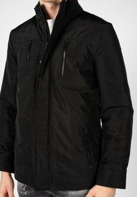 Giacca nera con colletto alto, tasche sul petto con zip, tasche laterali e cuciture distintive. Realizzata con un materiale liscio e leggero.